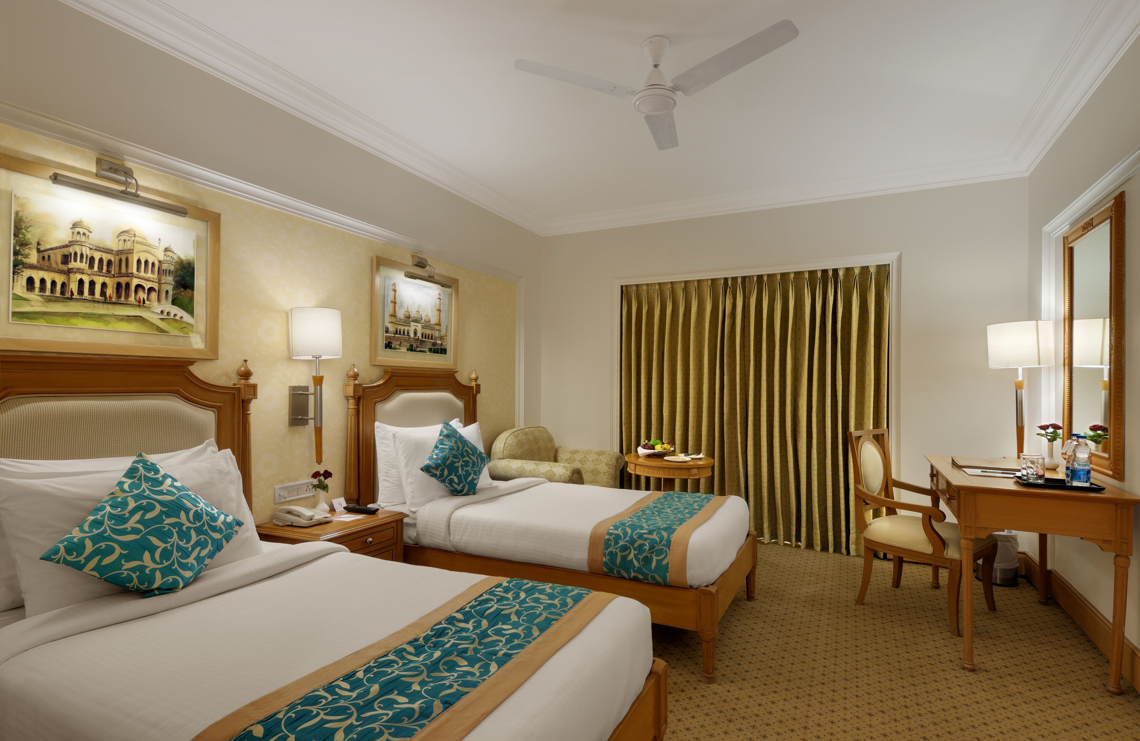 Quarto de hotel com duas camas, uma mesa, uma cadeira, luminária de mesa, telefone, pequena mesa com um vaso de flores e uma grande janela com cortinas.