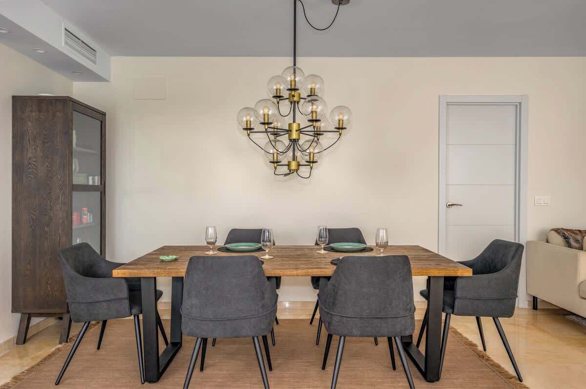 A dining area with a wooden table, four grey upholstered chairs, a chandelier, and a white door in the background.