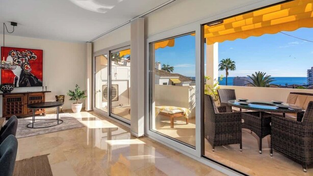 Una sala de estar moderna con un amplio ventanal corredera que da a un balcón con vista al mar.