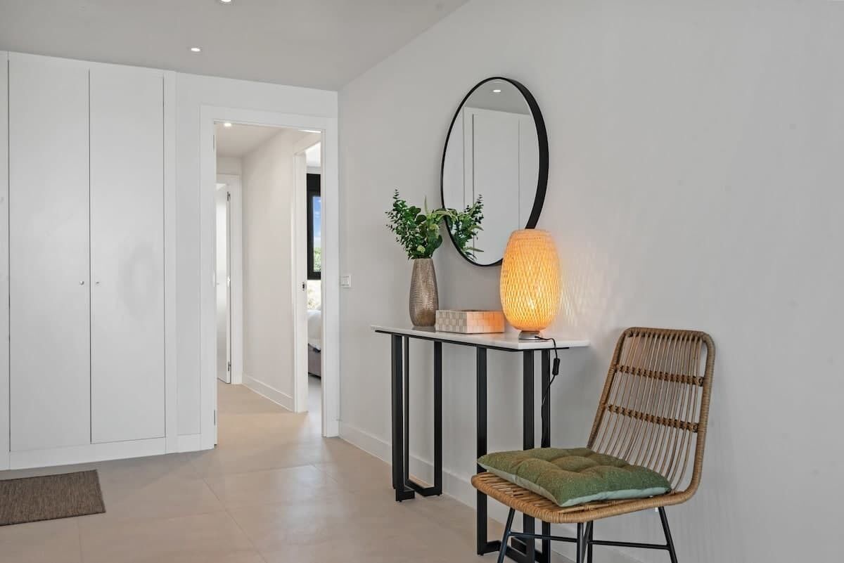 En gang med et hvidt skab, en fletstol med pude, et bord med lampe og et spejl på væggen.