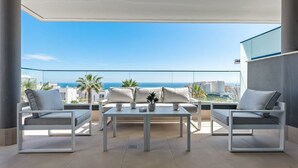Un balcón moderno al aire libre con un conjunto de sofá blanco, una mesa y vistas al océano y al horizonte de la ciudad.