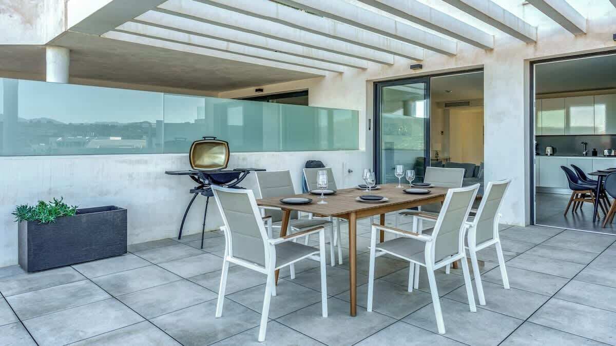 A modern outdoor dining area with a wooden table and white chairs, a black planter with green plants, and a glass partition wall.