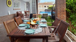 Una mesa de madera dispuesta para el desayuno, con sillas, una piscina al fondo y un muro de ladrillo.