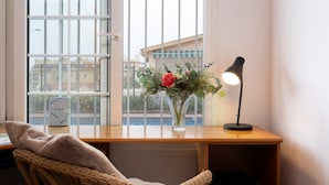 Una habitación con un escritorio, una silla de mimbre, un florero con flores y una lámpara.