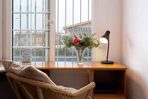 A room with a desk, a wicker chair, a vase with flowers, and a lamp.