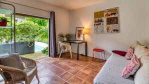 A room with a bed, a desk, a chair, a red stool, and a view of a swimming pool. - Oasis Centre Charme House with Shared Pool, Wi-Fi, and Air Conditioning (Montpellier)