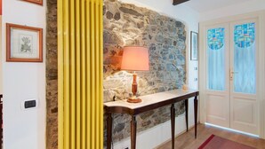 Un mur de pierre, un rideau jaune, une table avec une lampe, des tableaux encadrés et une porte avec des vitraux.