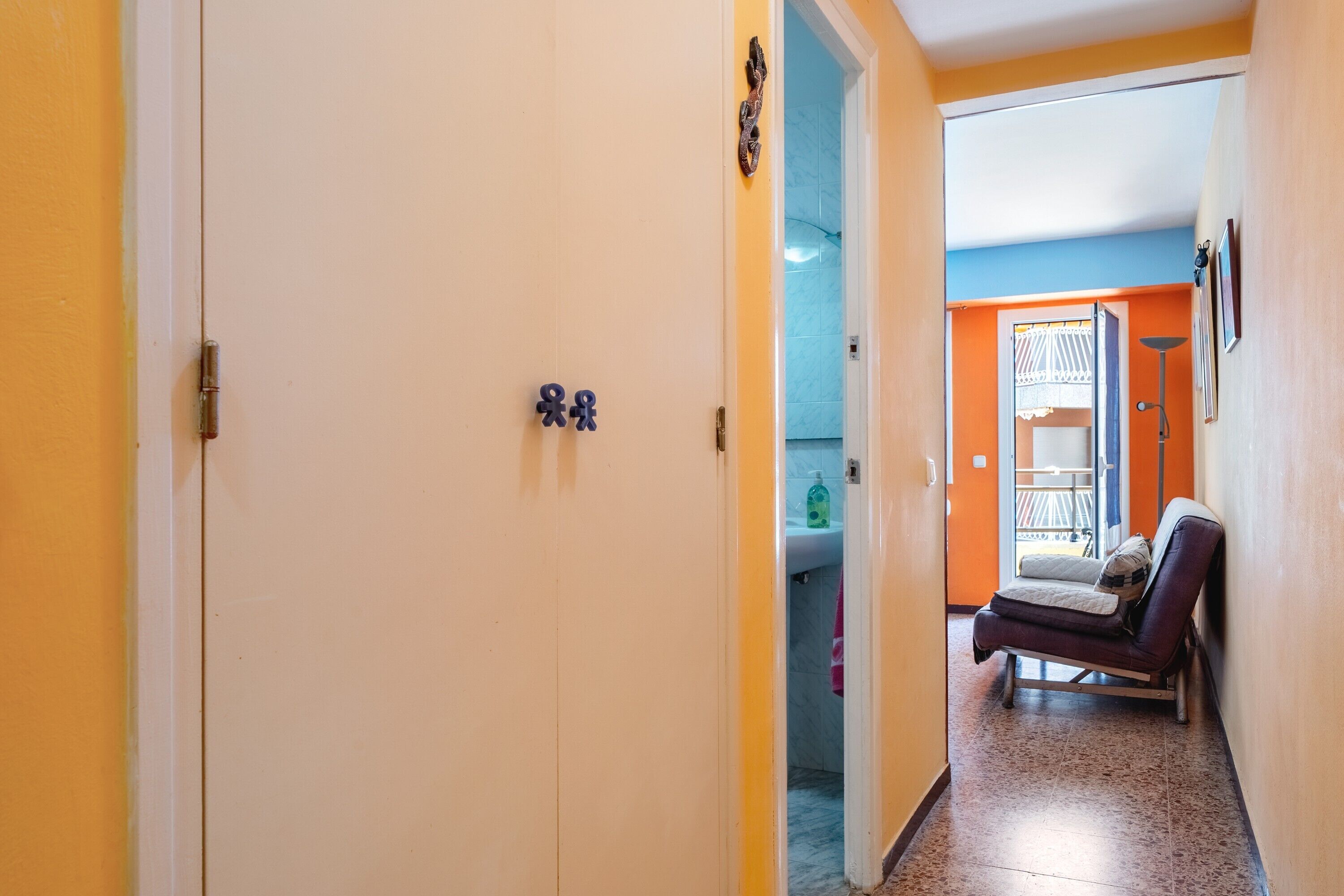 Un pasillo con una puerta, un banco y vista a un baño con lavabo e inodoro.