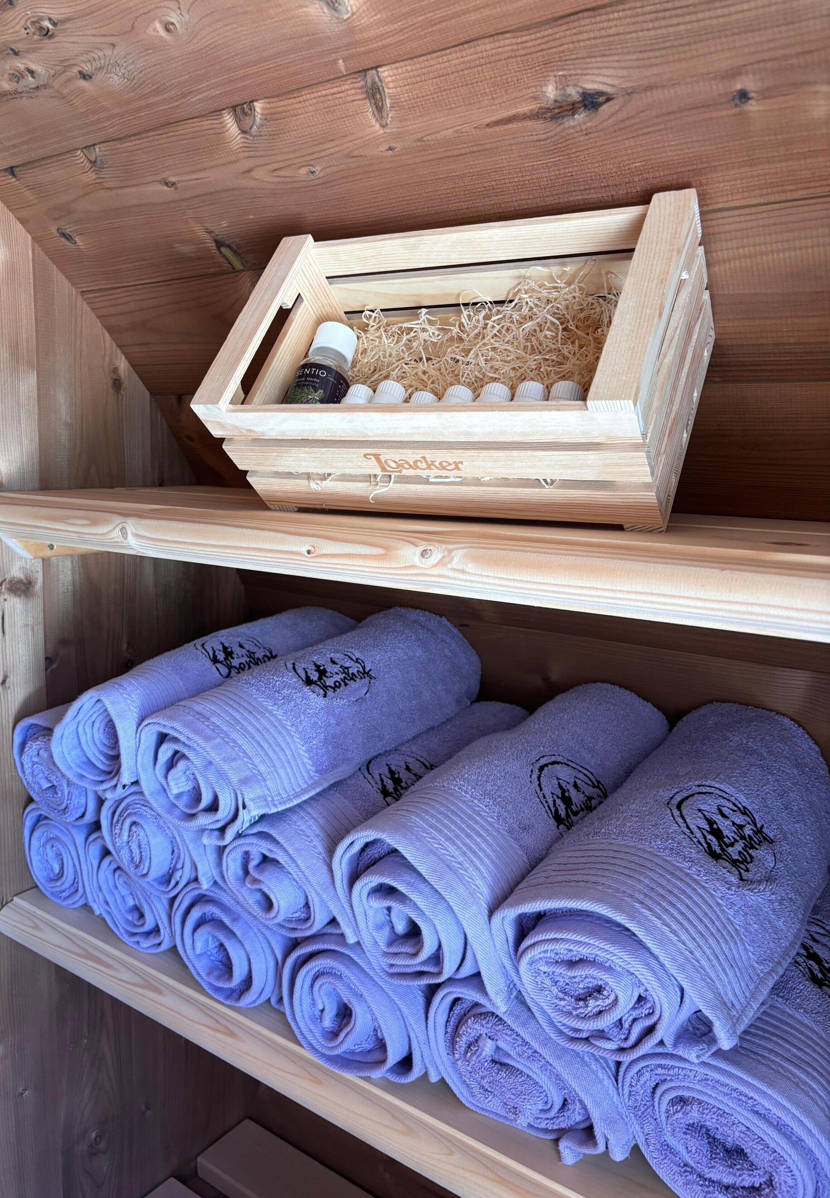 A wooden shelf with neatly rolled lavender towels and a wooden box containing a bottle and a small container.