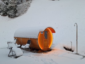 A wooden cabin with a snow-covered roof, a door illuminated from the inside, and a metal chair outside.
