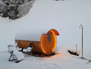 Una cabaña de madera con techo nevado, una puerta iluminada desde el interior y una silla de metal en el exterior.