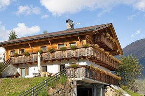 A chalet with wooden balconies, a stone staircase, and a mountain backdrop.