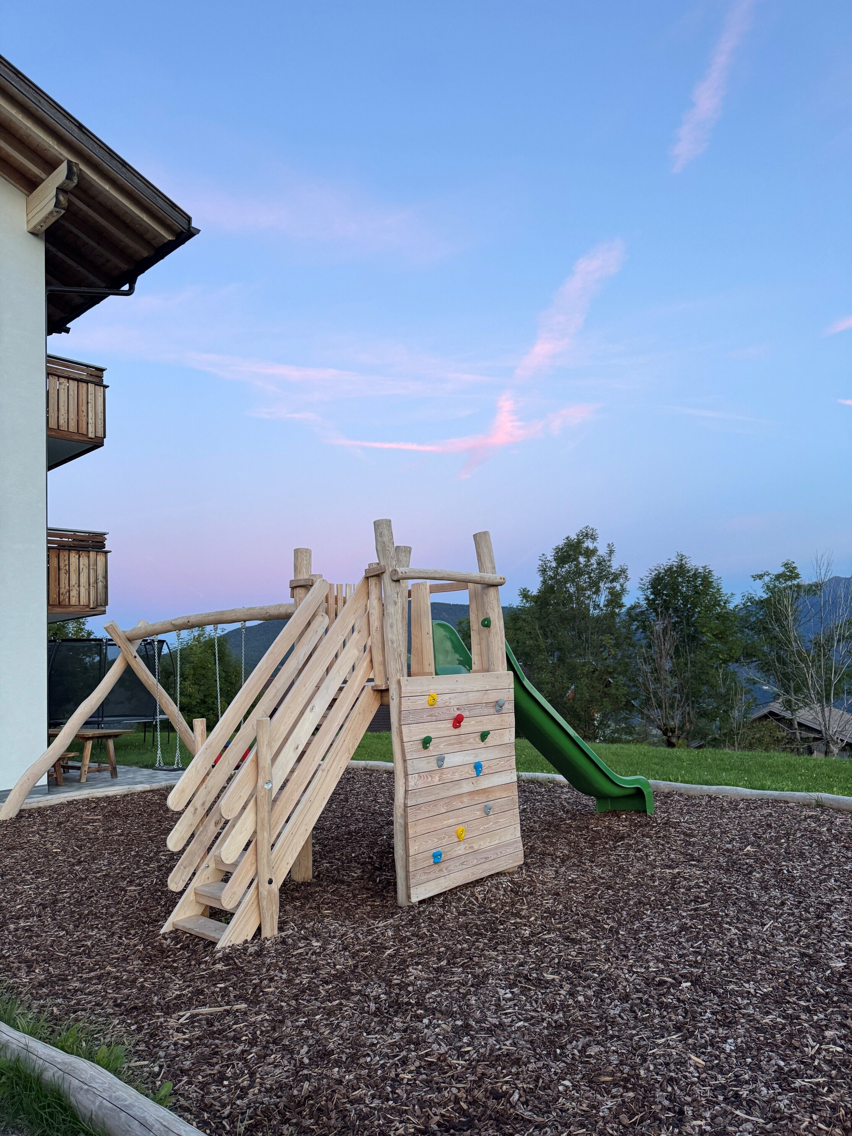 Ein hölzerner Spielplatz mit Rutsche und Kletterwand, umgeben von Rindenmulch und Bäumen.