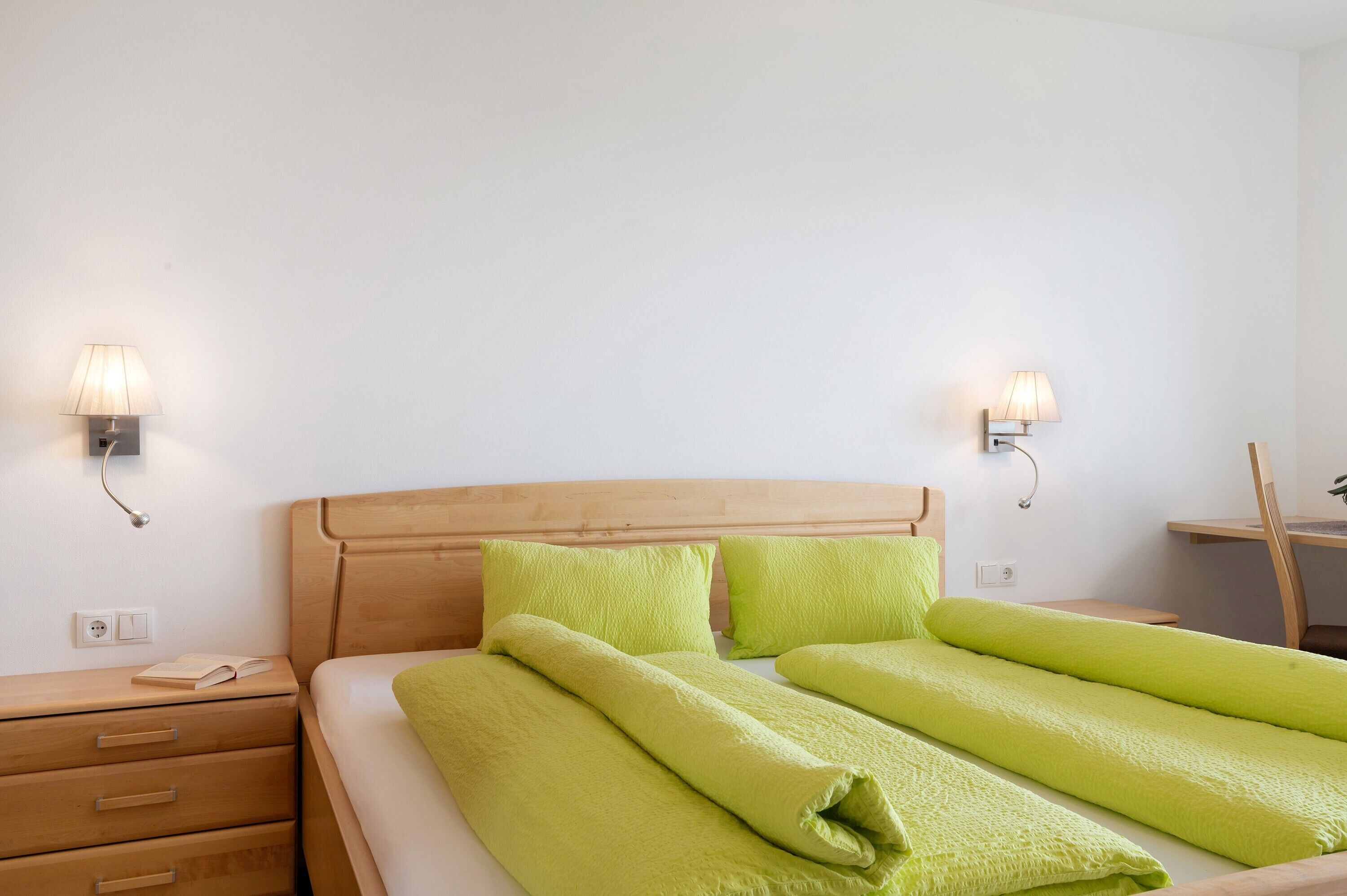 Habitación de hotel con cabecera de madera, dos lámparas de noche, mesita de noche de madera, cama con ropa de cama verde y un escritorio con silla.