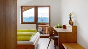 Een kamer met een bed, een bureau, een stoel, een houten bankje, een raam met uitzicht op bergen en een kleine plant op het bureau.