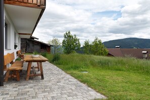 Eine Terrasse mit Holztisch und Bänken, ein Pflasterweg und Blick auf einen Berg.