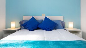 Una cama con ropa de cama blanca y almohadas azules, dos mesitas de noche con lámparas y un interruptor de luz en la pared.