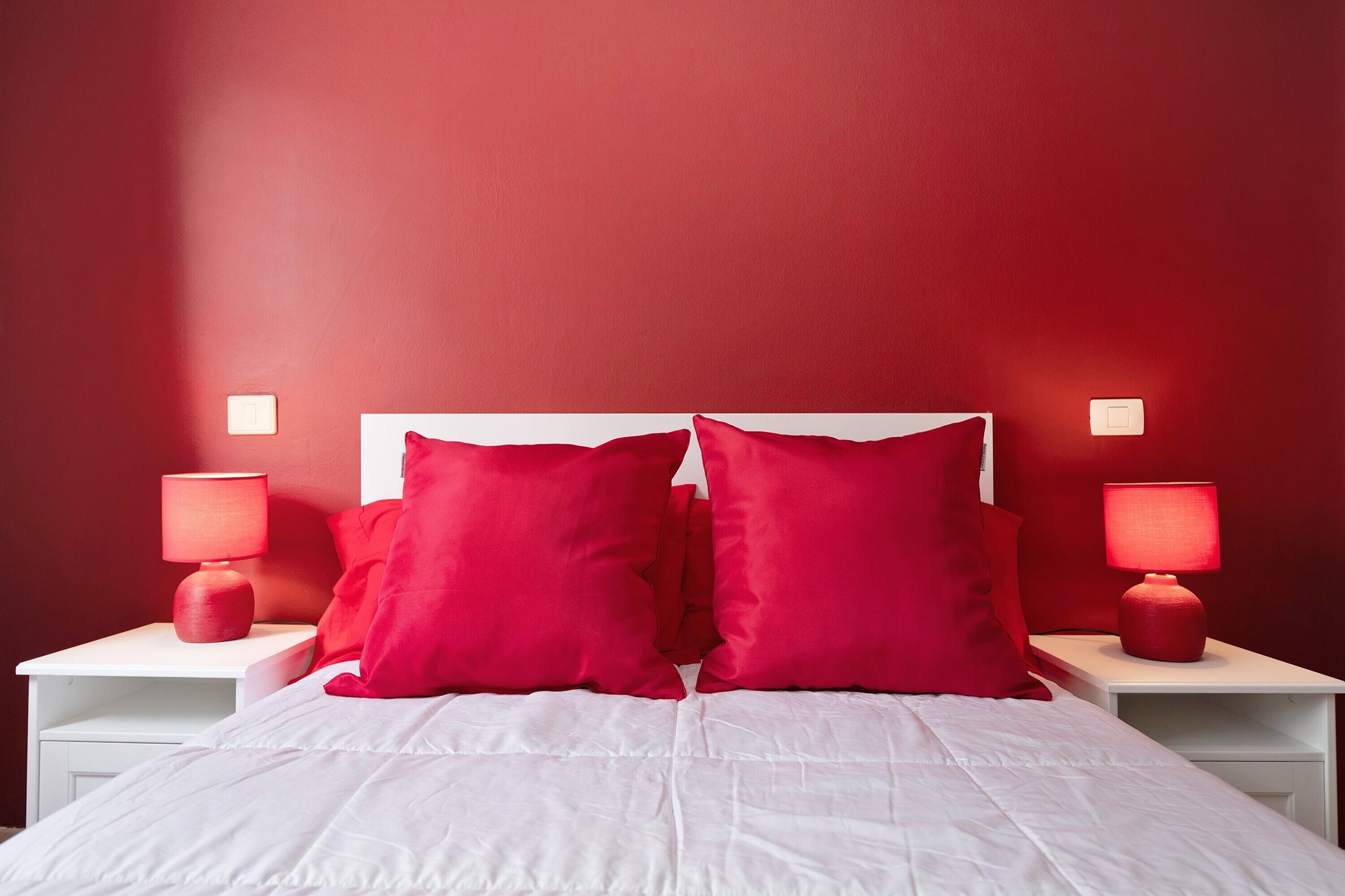 A bed with white bedding and red pillows, two bedside tables with lamps, and a red wall.