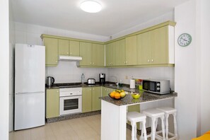 Une cuisine de taille réduite avec des meubles verts, un réfrigérateur blanc, un four à micro-ondes, un évier et un panier de fruits posé sur le plan de travail.
