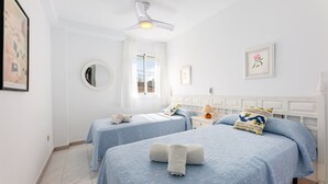 Une chambre d’hôtel avec deux lits, un ventilateur de plafond, une fenêtre avec des rideaux, une table de chevet avec une lampe et un tableau encadré au mur.