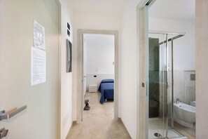 A narrow hallway leading to a bedroom with a blue bedspread, a glass-enclosed shower, and a visible toilet.
