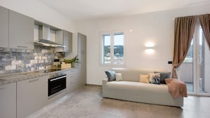 Una cocina moderna con armarios grises, horno empotrado y salpicadero de piedra. Hay un sofá color beige con cojines y vistas al exterior a través de una ventana.
