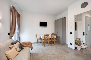 A modern living room with a sofa, a dining table with chairs, a flat-screen TV, and a door leading to another room.
