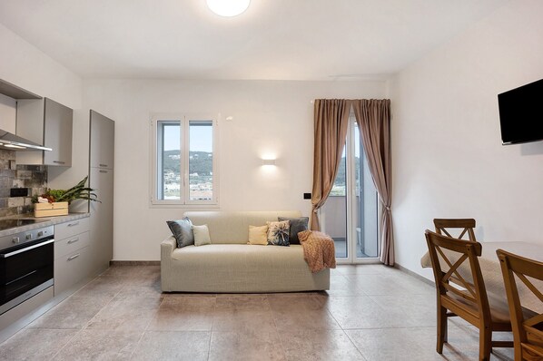 A modern kitchen with a built-in oven, a dining area with a wooden table and chairs, a sofa with cushions, and a large window with curtains.