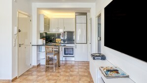Cuina compacta amb armaris blancs, una taula de dinar amb cadires, forn integrat i un televisor de pantalla plana a la paret.