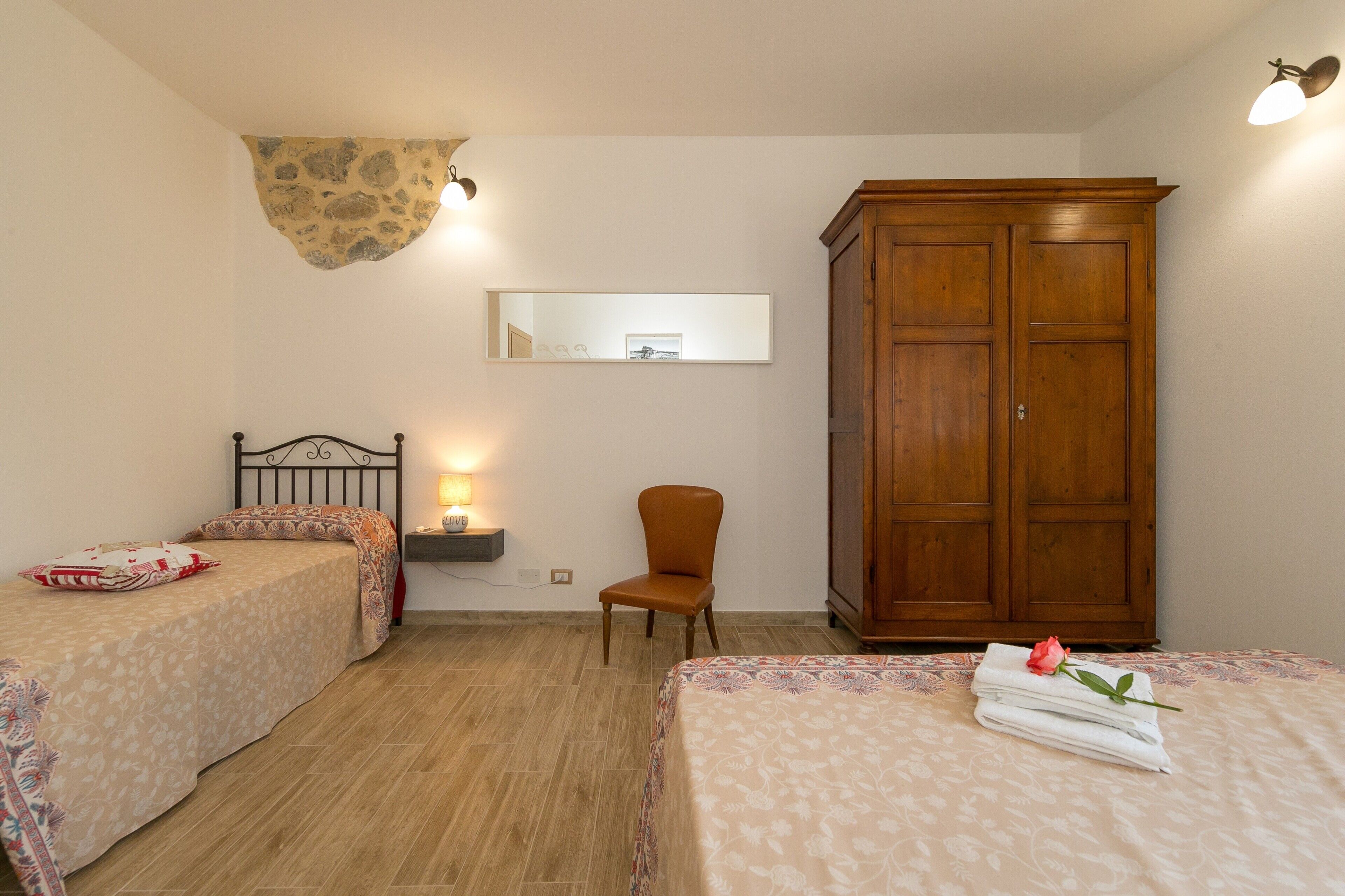 A bedroom with two beds, a wooden wardrobe, a chair, a mirror, and a wall-mounted light fixture.