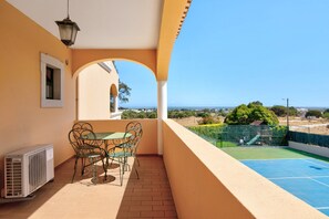 A balcony with a table and chairs, a view of the sea, and a tennis court.