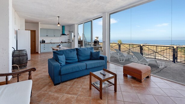 Un amplio salón con un sofá azul, una mesa de centro de madera y vistas al mar a través de grandes ventanales.