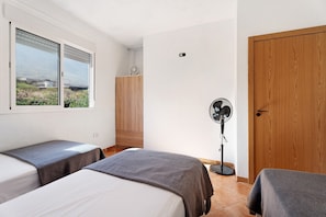 Une chambre avec deux lits, une armoire en bois, un ventilateur sur pied et une fenêtre donnant sur un beau paysage.