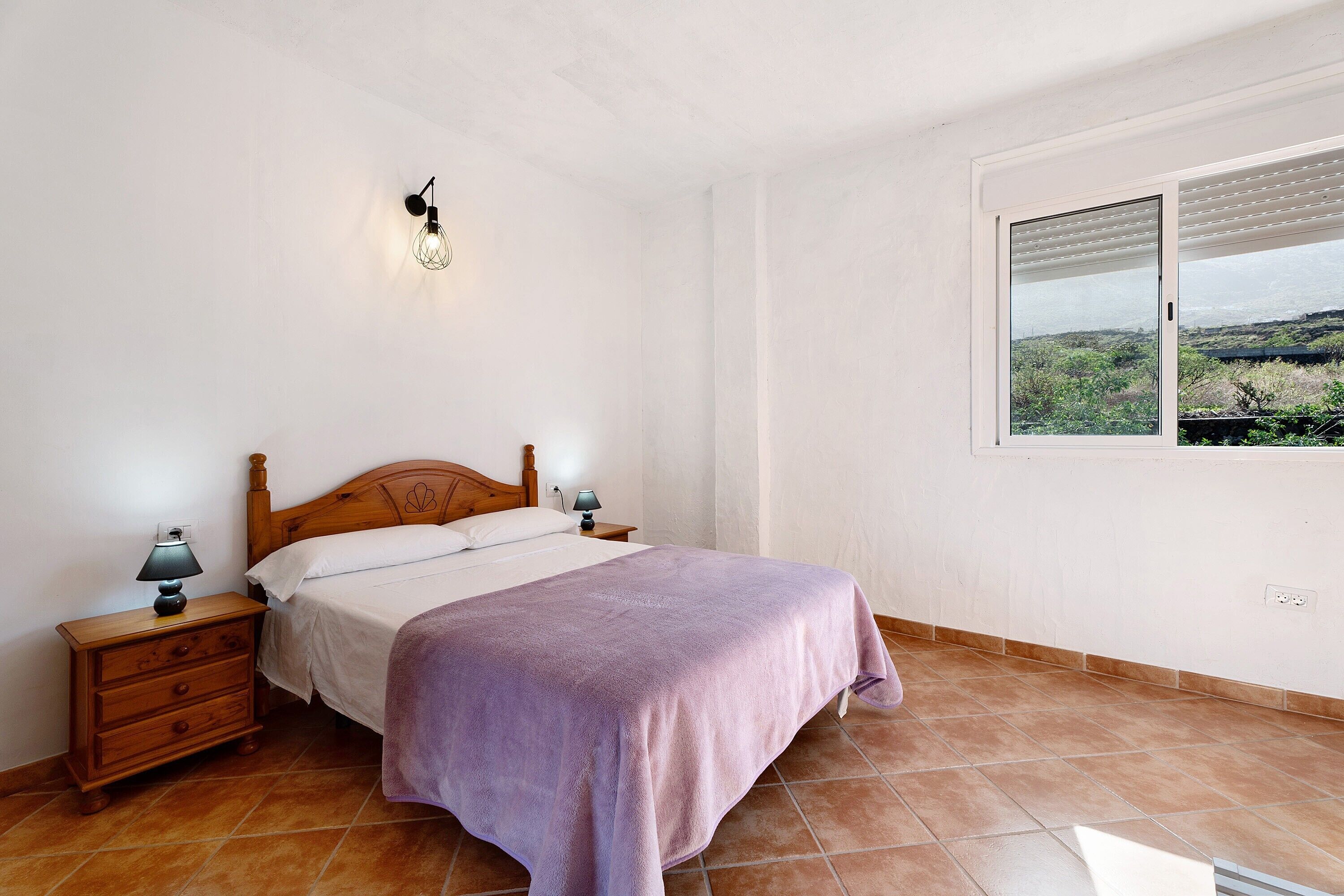 A bedroom with a bed, nightstands, a wall-mounted lamp, and a window with a view.