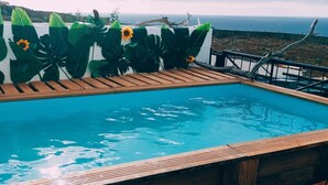 Una piscina con cubierta de madera, rodeada de un muro decorativo con hojas tropicales, y vista al océano.