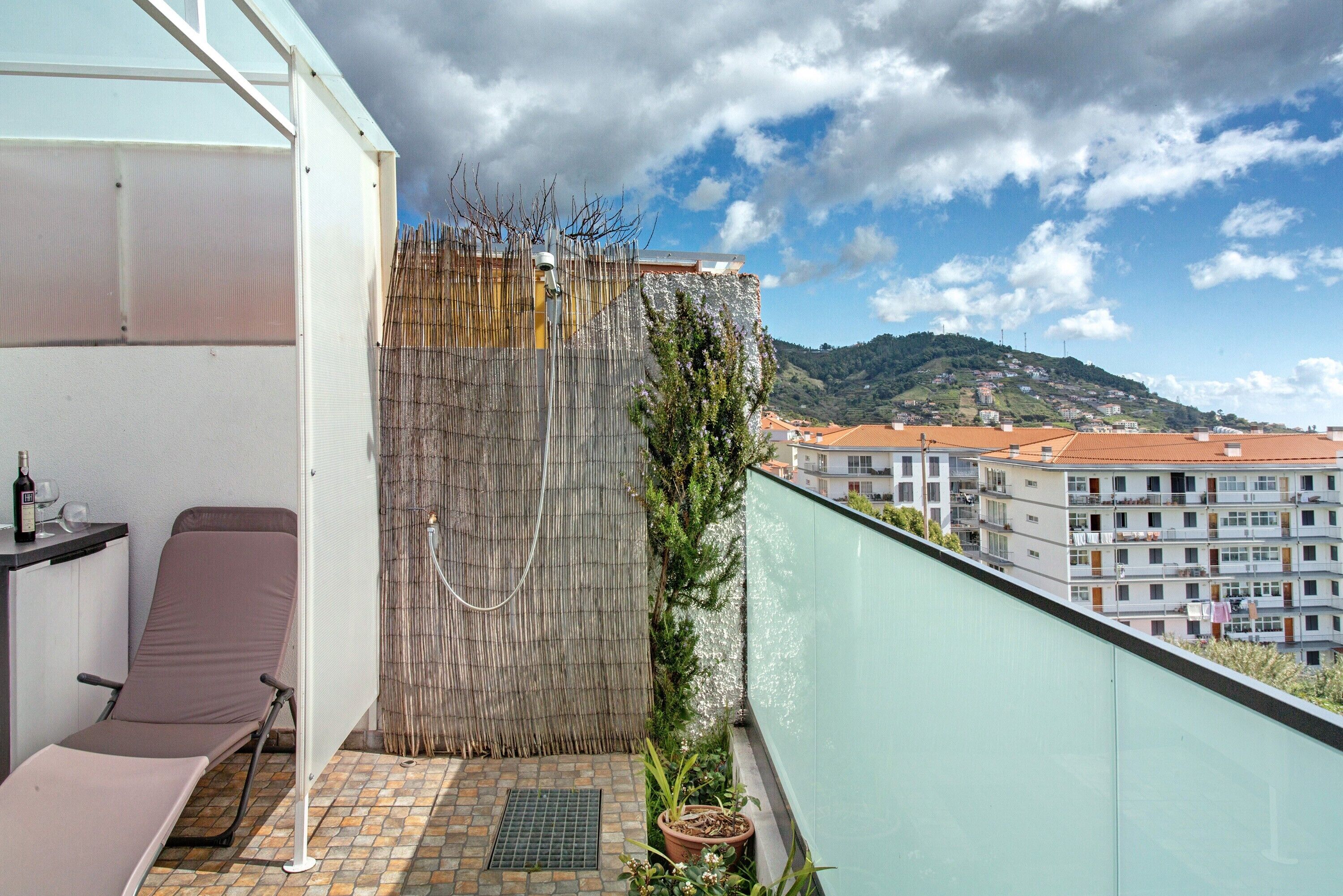 A balcony with a chair, a potted plant, and a view of buildings and hills.