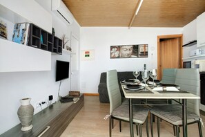 Un área de comedor moderna con una mesa preparada para cuatro personas, un televisor montado en la pared y una cocina visible a través de una puerta abierta.