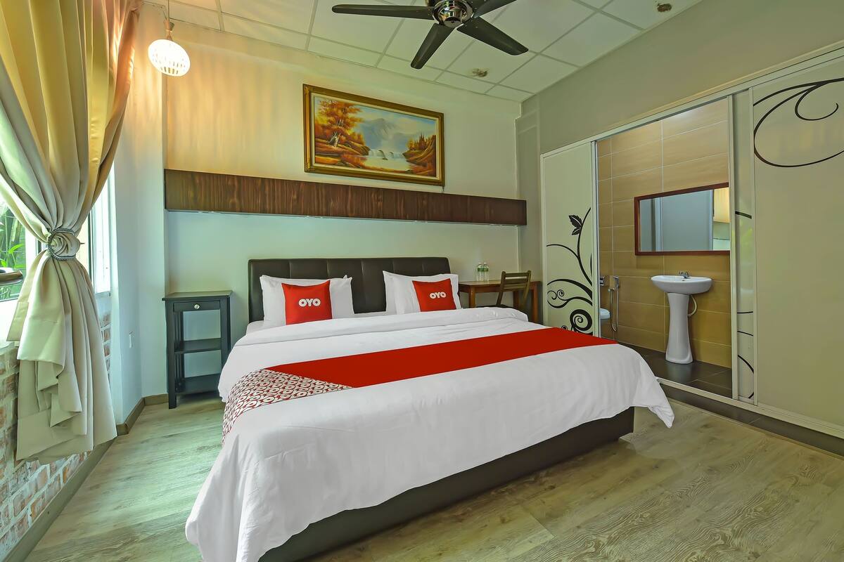 A hotel room with a large bed, a ceiling fan, a painting on the wall, a bedside table, and a bathroom with a sink and mirror.