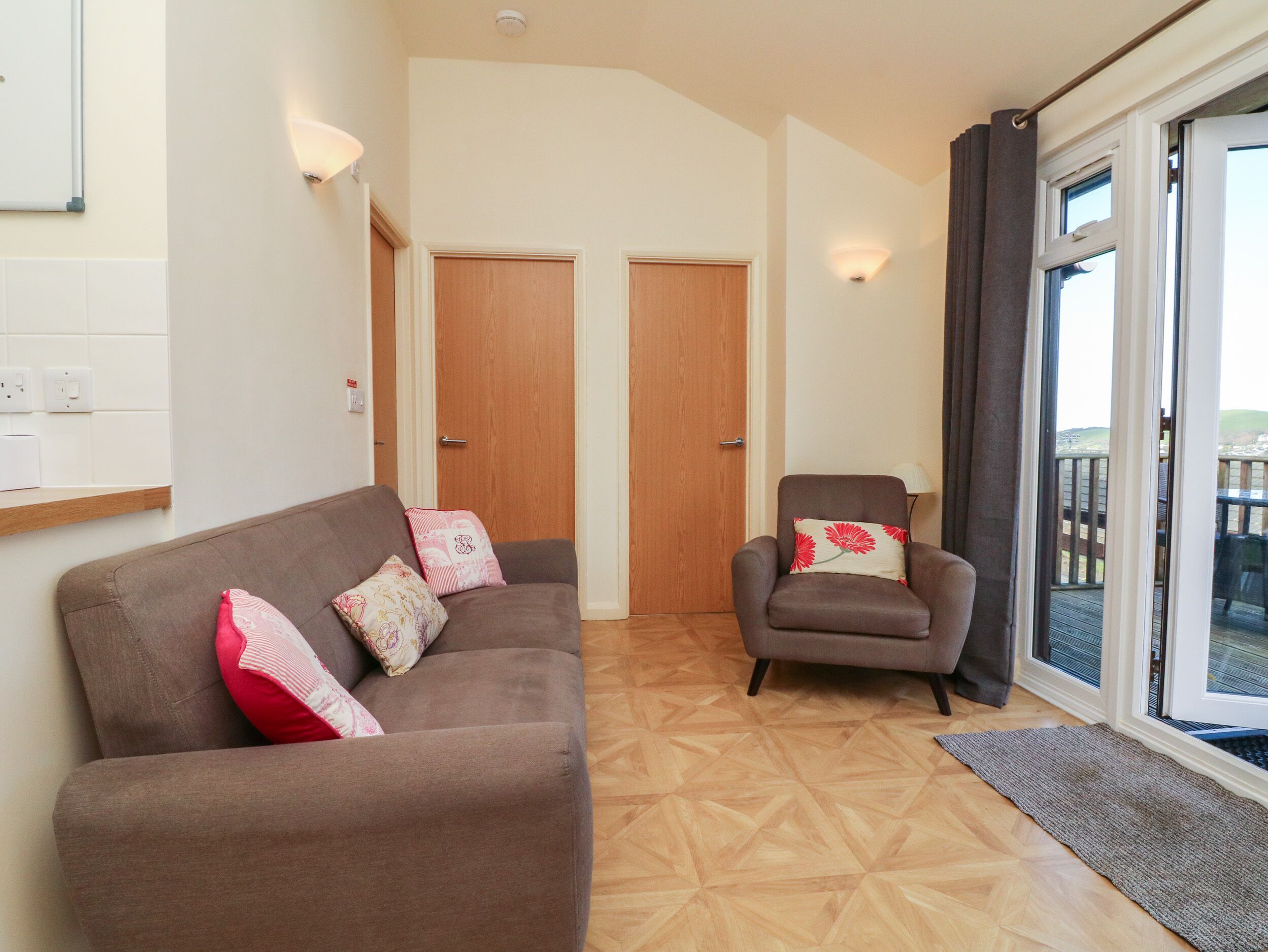 Ein Wohnzimmer mit braunem Sofa, einem braunen Sessel und Holzboden. Es gibt zwei Türen und ein Fenster mit Vorhängen.