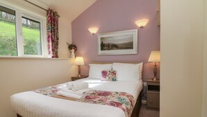 A neatly made bed with white linens and a floral-patterned bedspread, two bedside lamps, a framed picture on the wall, and a window with curtains. - 12 Manleigh Park (Ilfracombe)