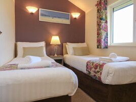Dos camas individuales con sábanas blancas, una mesita de noche con lámpara, aire acondicionado instalado en la pared y una ventana con cortinas florales.