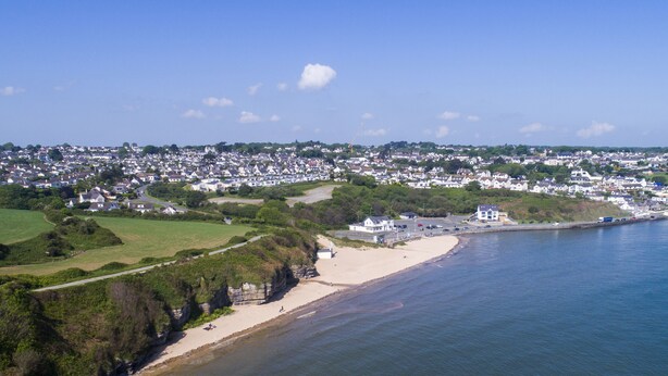 A coastal town with a sandy beach, a rocky shoreline, and residential buildings in the background. - Tinkers Patch (Tyn-y-Gongl)