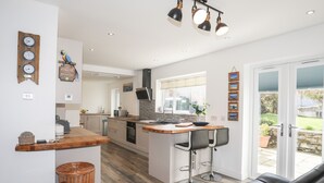 A modern kitchen with a central island, bar stools, and a dining area with a black leather couch. - Tinkers Patch (Tyn-y-Gongl)