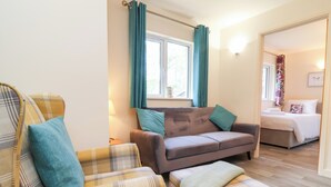 Een gezellige woonkamer met een bank, fauteuil en poef, een raam met gordijnen, en een bed in de aangrenzende kamer.