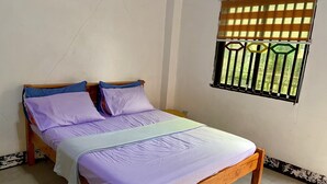 Ein Einzelbett mit Holzrahmen und weißer Matratze, ein Fenster mit Metallgitter und eine weiße Wand mit schwarz-weiß gestrichenem Sockel.