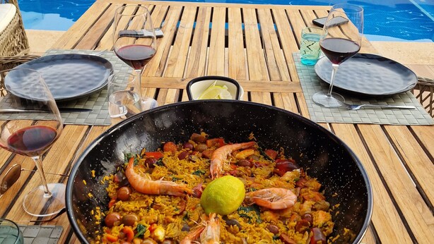 Ein Esstisch mit einer großen Paella-Pfanne, einem Weinglas und einem Pool im Hintergrund.