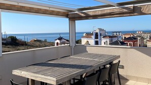 Eine Terrasse mit Holztisch und -stühlen, einer Pergola und Blick auf das Meer und die Gebäude.