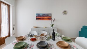 Ruokapöytä on katettu lautasilla, kulhoilla, laseilla ja siinä on myös pullo. Seinällä on auringonlaskua kuvaava taulu ja huoneessa on myös kello.