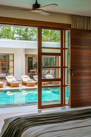 A bedroom with a large bed, a view of a swimming pool, and a sliding glass door leading to a balcony with lounge chairs. - Villa KClub River Front 4 (Canggu)
