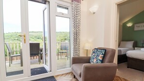 A room with a wooden herringbone floor, a brown armchair with a floral pillow, a bed with white bedding, and a view of greenery through a sliding glass door. - 14 Manleigh Park (Ilfracombe)